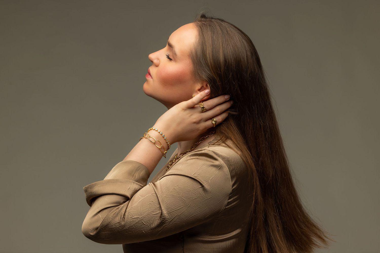 Woman in beige jacket posing sideways with hands on neck, wearing gold bracelets against gray background.