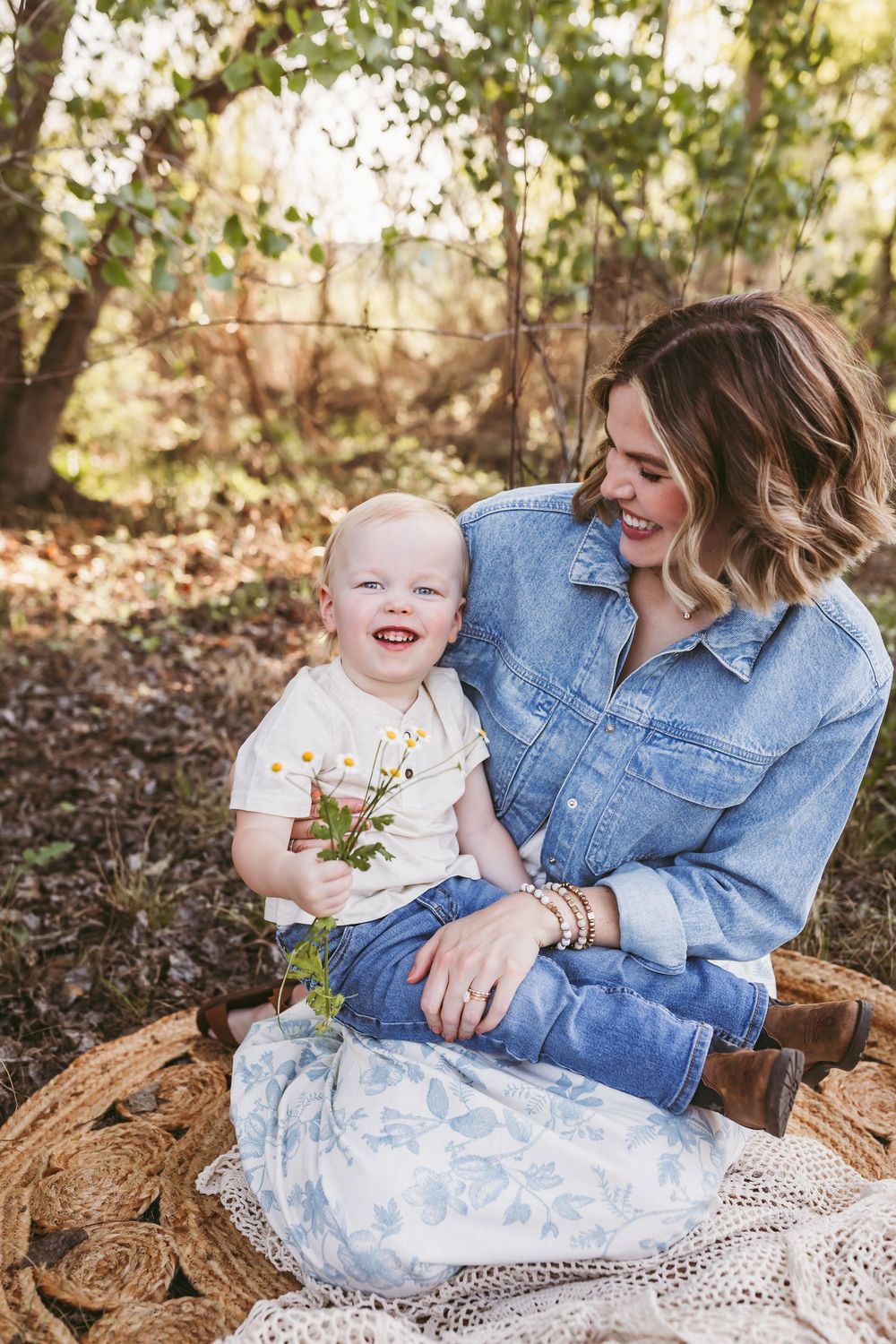 A laughing mother sits outdoors on a wicker seat holding her happy toddler who clutches wildflowers near beaumont ca
