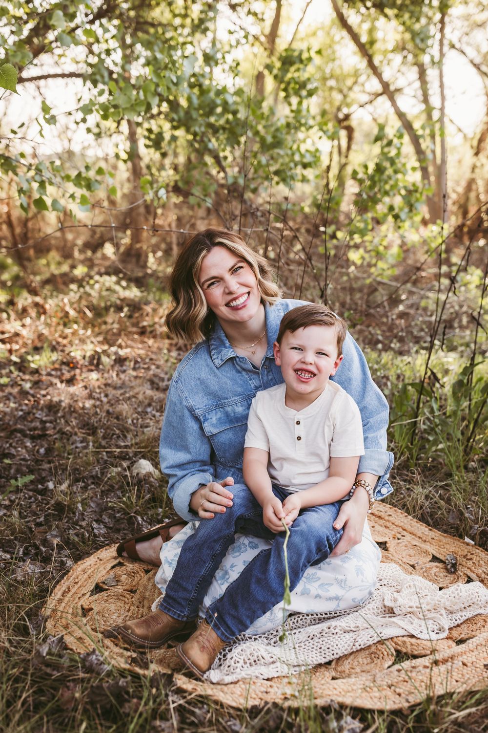 A smiling mother and toddler son sit together on a woven mat outdoors, surrounded by lush green trees close to yucaipa ca