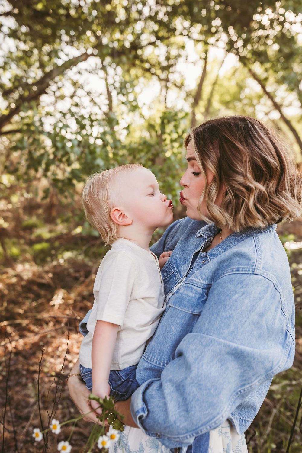 A mother holds her toddler son as they share a sweet kiss outdoors among trees and wildflowers with heidi grace photography