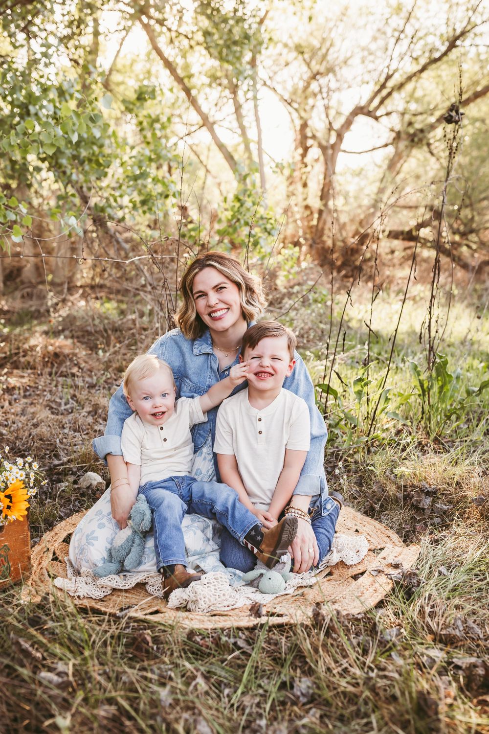 A smiling mother sits outdoors on a blanket with her two young boys in a sunlit wooded setting.