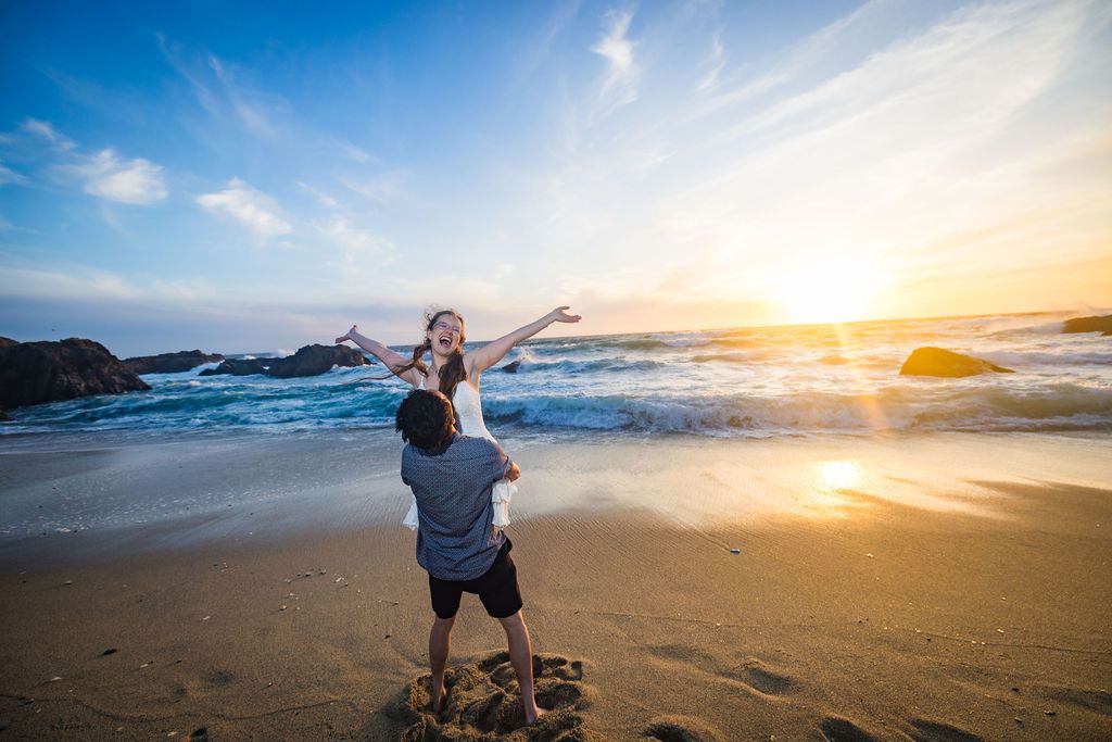 Couple enjoying a romantic sunset on a sandy beach, man lifting woman with ocean waves and golden sky in the background.