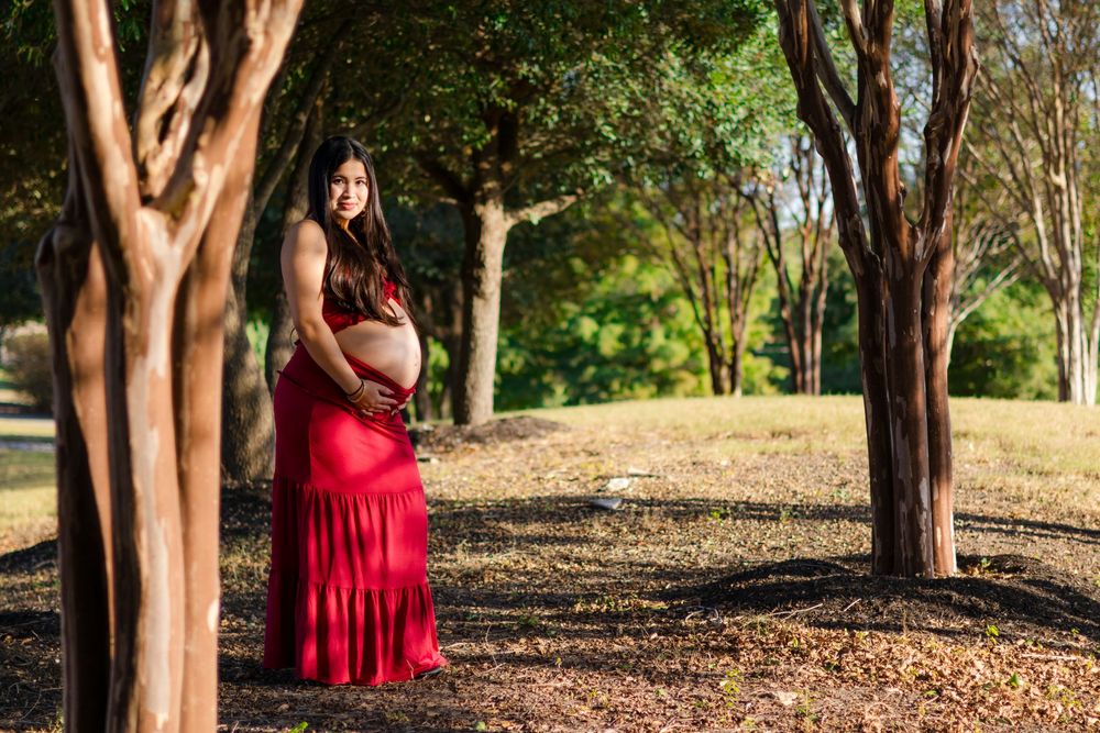 An editorial maternity portrait of a woman in a red gown standing among tall trees in a sun-drenched Austin park.