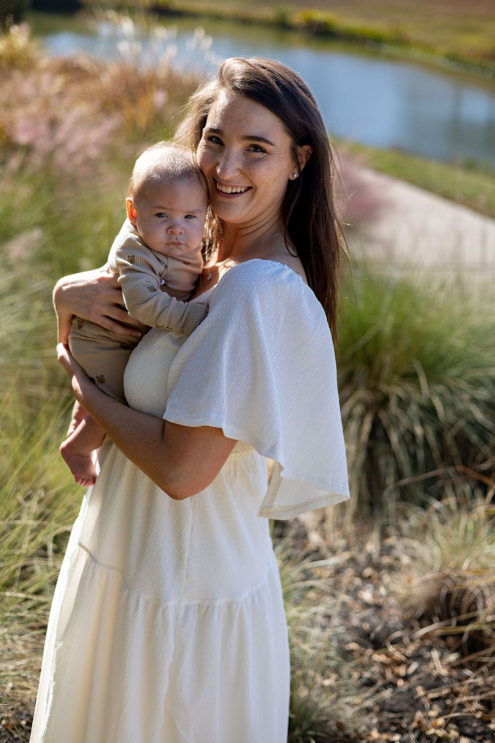 A beautiful outdoor motherhood portrait of a mother in a white dress holding her baby in a sunlit field near Austin, Texas.