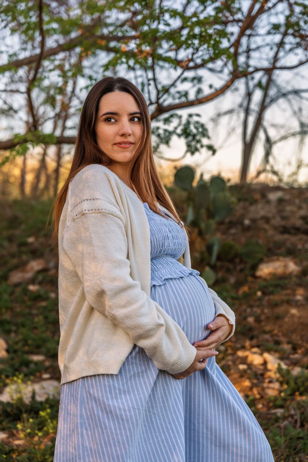 A natural light maternity portrait of a woman in a blue dress and cardigan, highlighting the un-staged nuances of her motherhood journey.