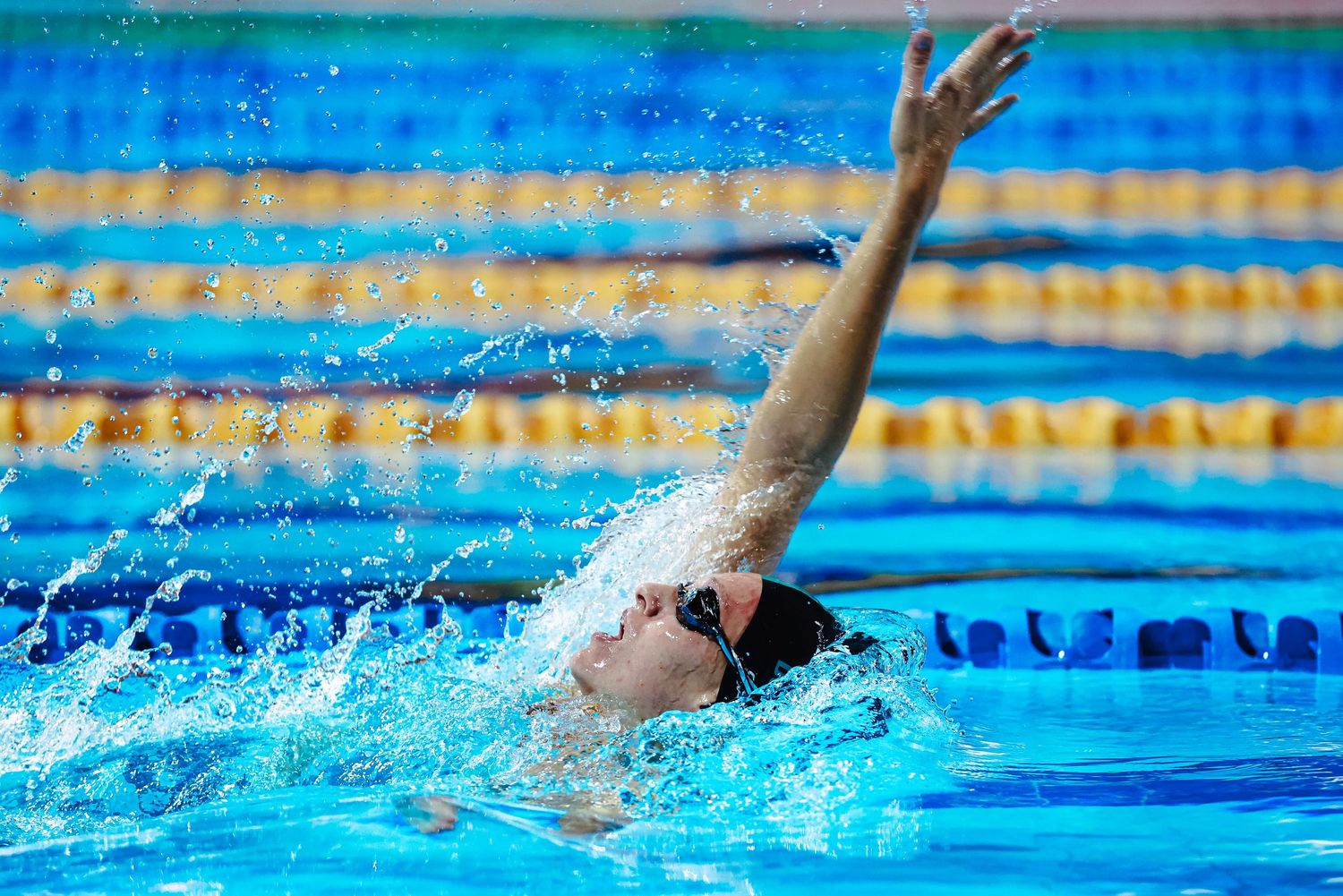 Competitive swimmer in red cap performs butterfly stroke, splashing water in a blue outdoor pool.