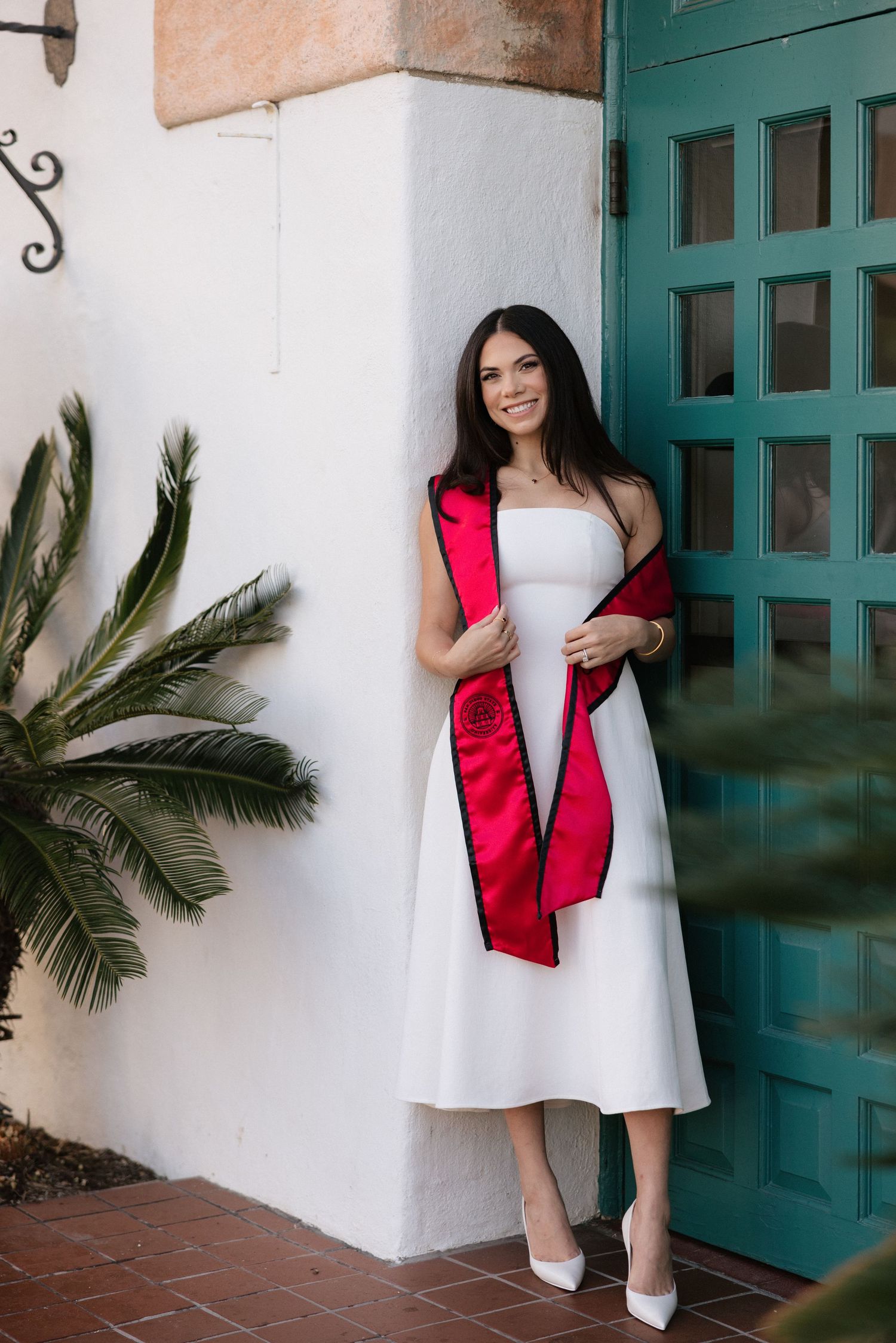Young woman in white midi dress and red graduation sash smiles beside teal door with tropical palm.
