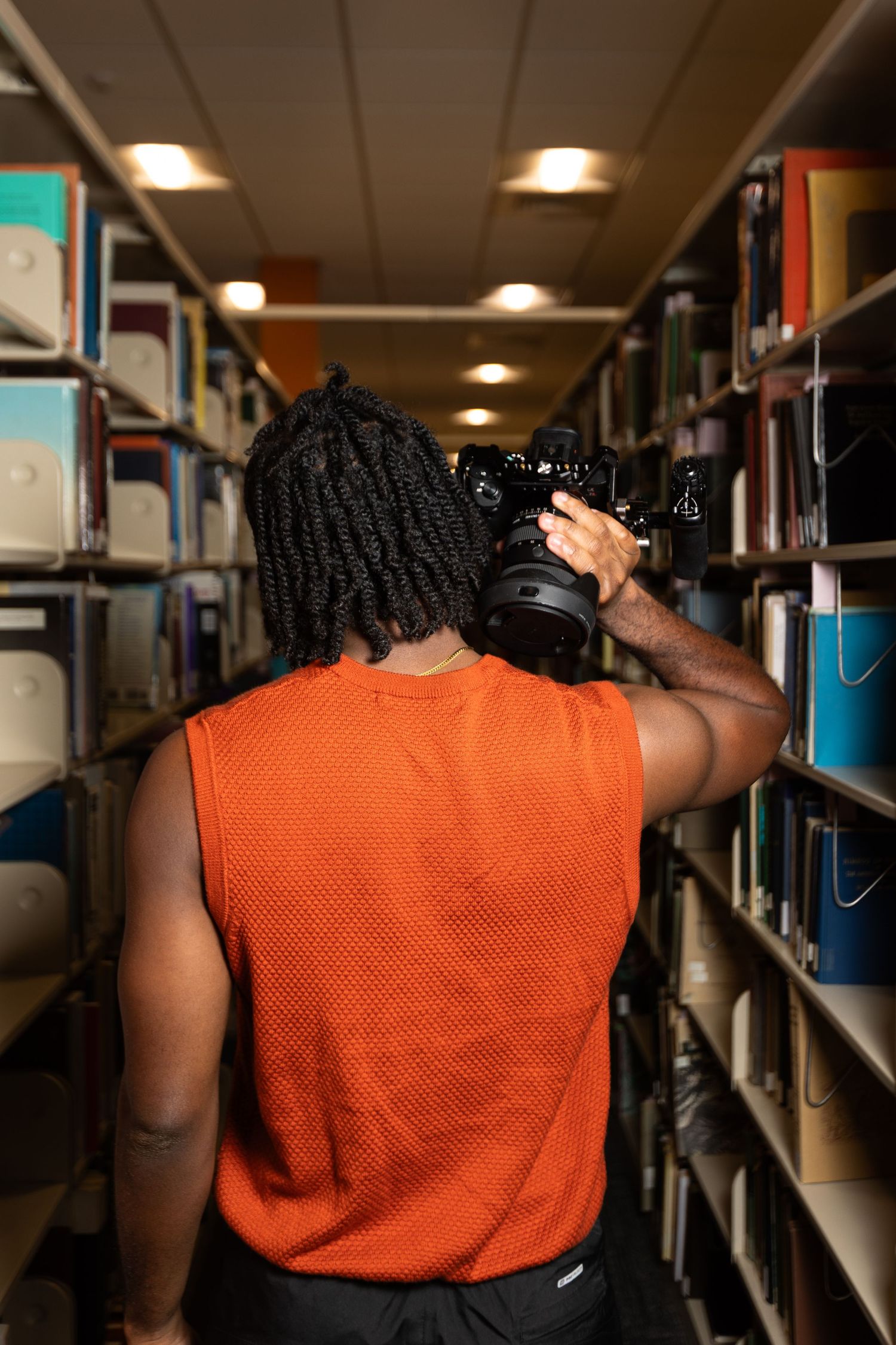 Person in orange sleeveless shirt browses through library bookshelves in dimly lit setting.
