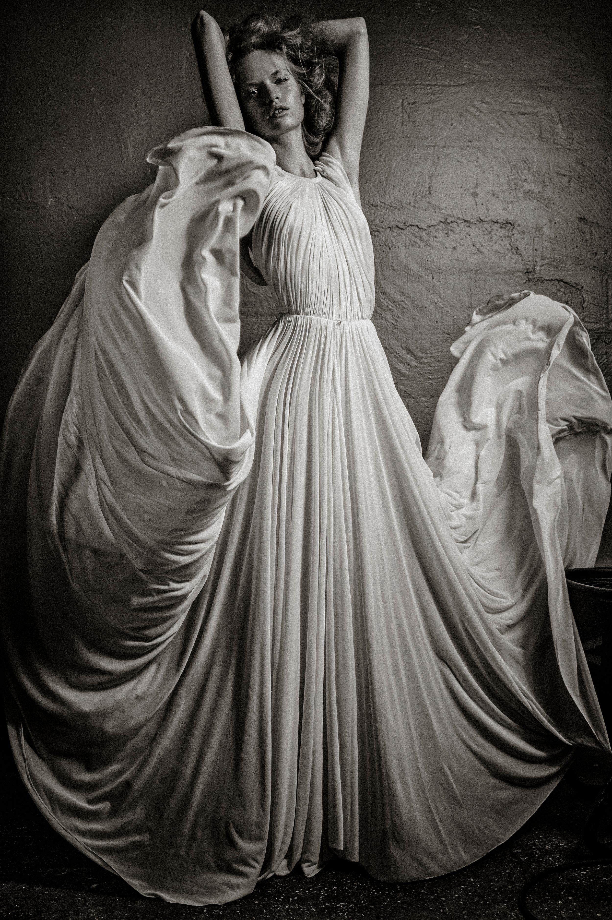 Black and white photo of a woman in a flowing white pleated gown with arms raised, fabric billowing dramatically.