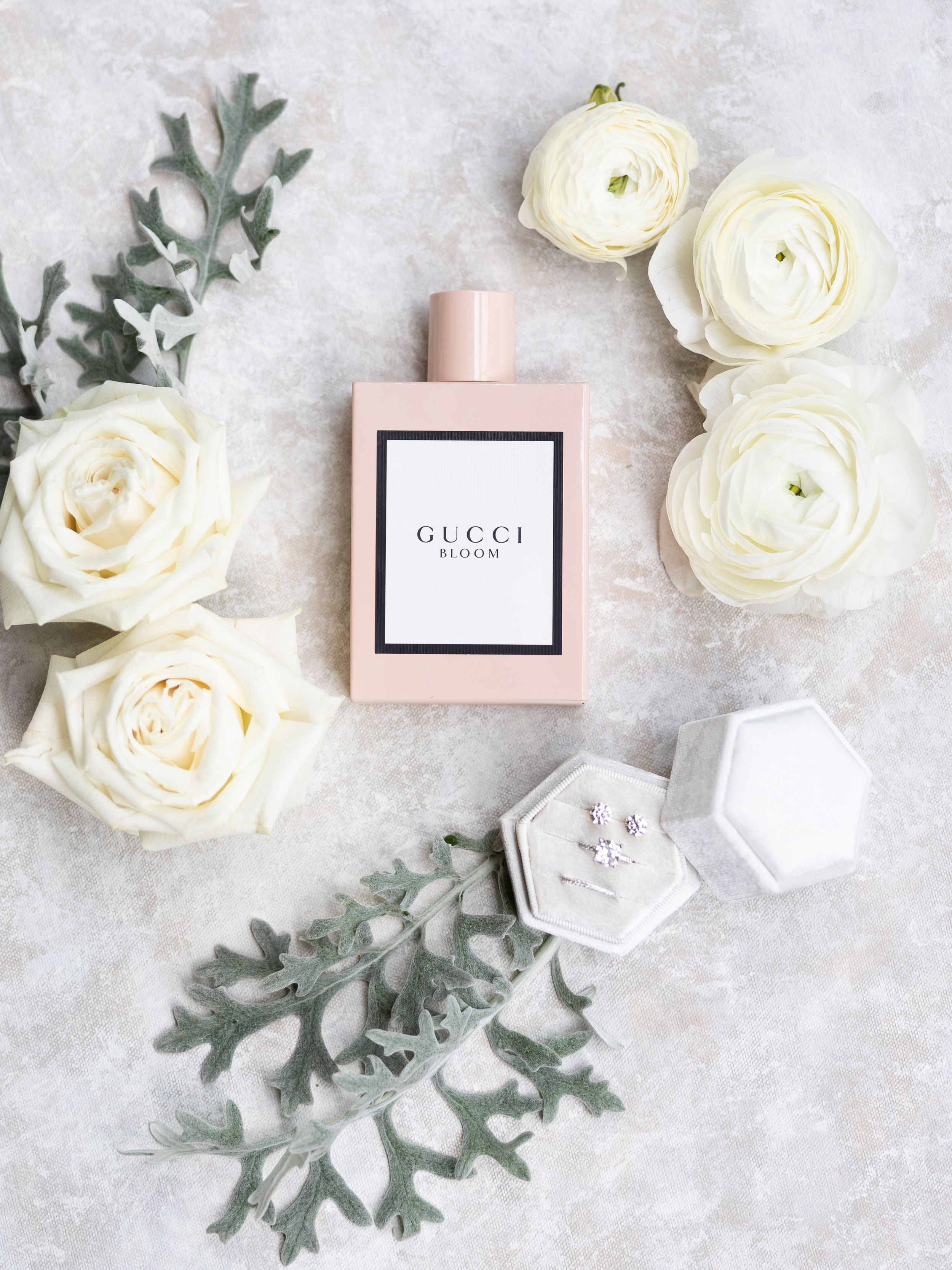 Gucci Bloom perfume bottle surrounded by white ranunculus roses and dusty miller with a hexagon ring box on marble.
