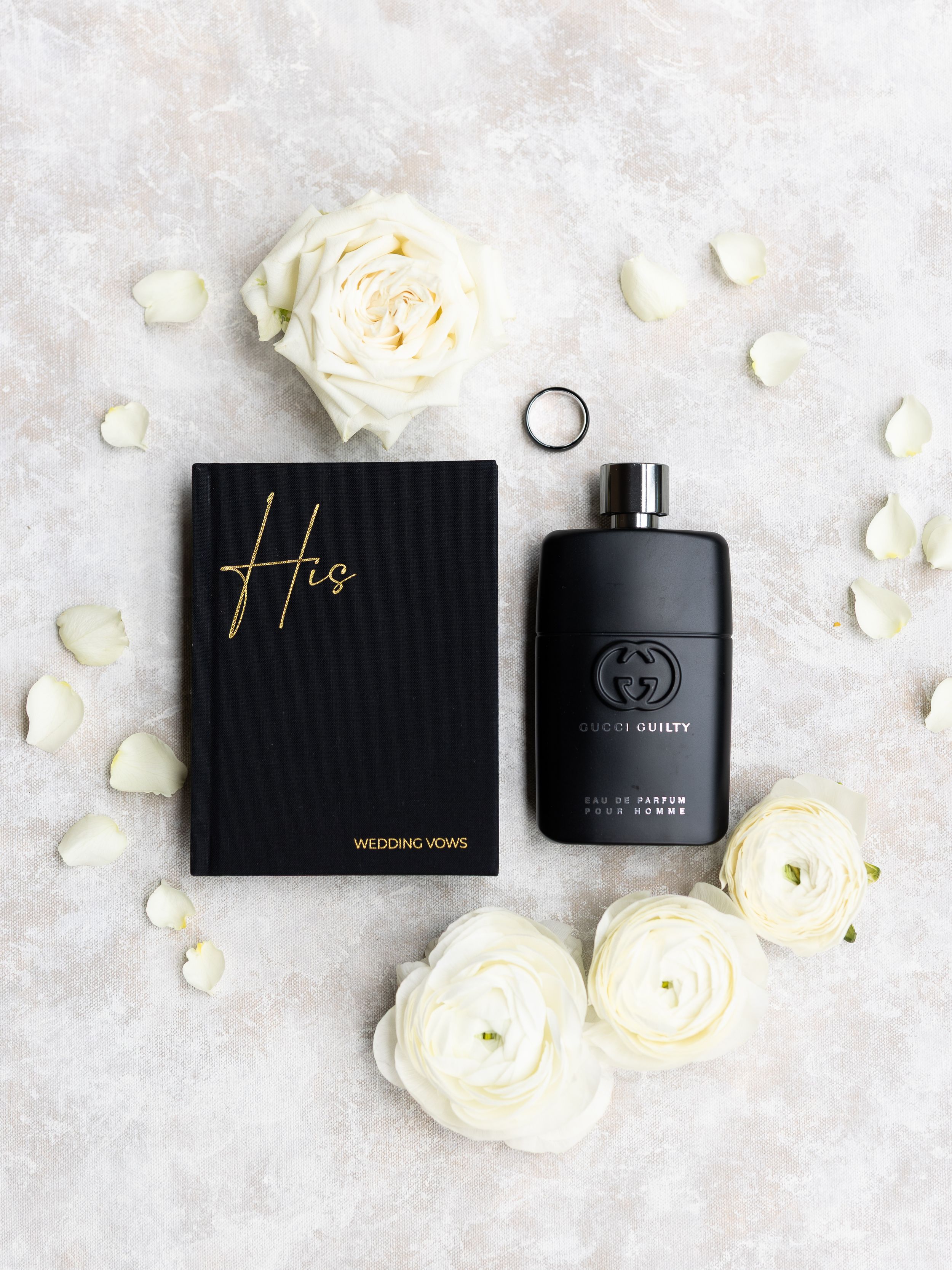 Gucci Guilty pour homme cologne beside a black wedding vows book with white roses and a ring on marble.