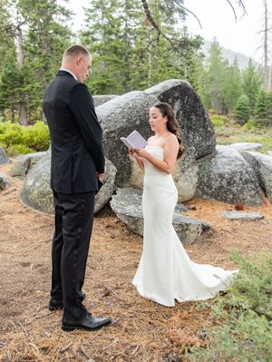 Bride reads vows to groom during outdoor wedding ceremony surrounded by granite boulders and pine trees.