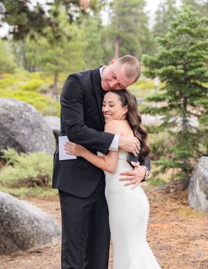 Bride and groom share a tender embrace outdoors among pine trees and boulders during their wedding day.