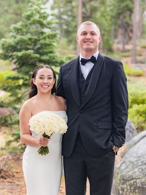 Bride in strapless white gown holding rose bouquet stands with groom in black tuxedo outdoors.