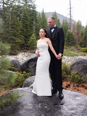 Bride and groom in formal attire standing on rocks surrounded by pine trees and mountains.