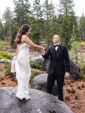 Bride and groom in formal wedding attire holding hands among pine trees and granite boulders in Tahoe forest.