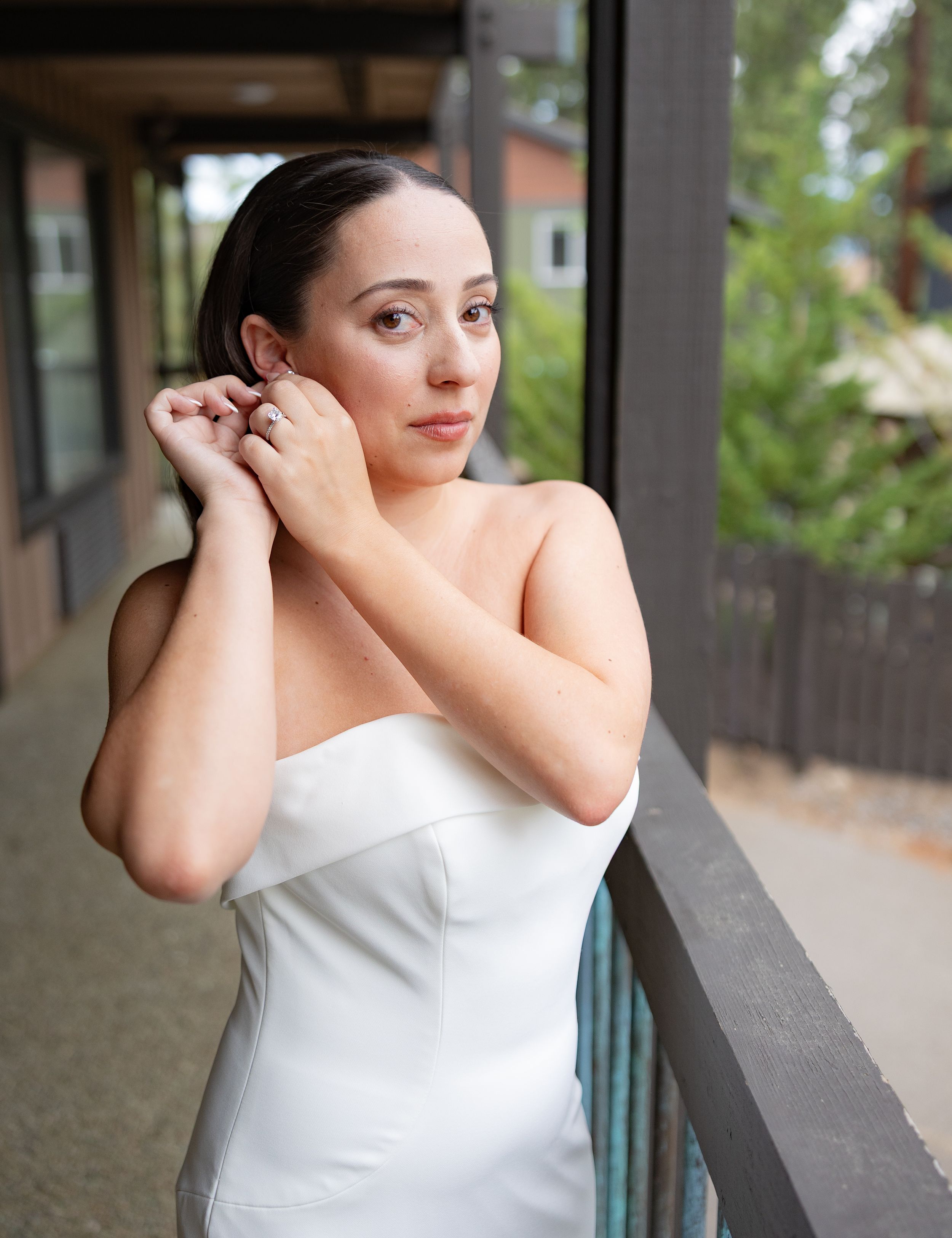 A bride in a white strapless gown adjusts her earring on a balcony with lush greenery in the background.