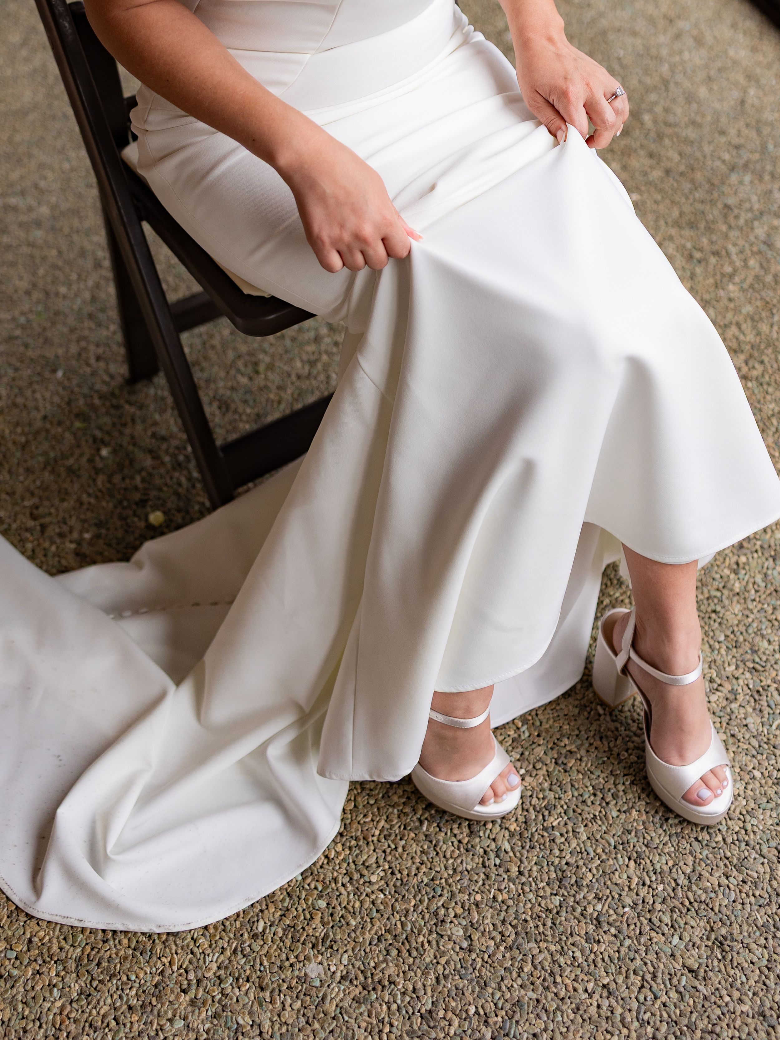 Bride in white satin wedding gown adjusting her skirt while seated on a wooden chair, wearing white heeled sandals.