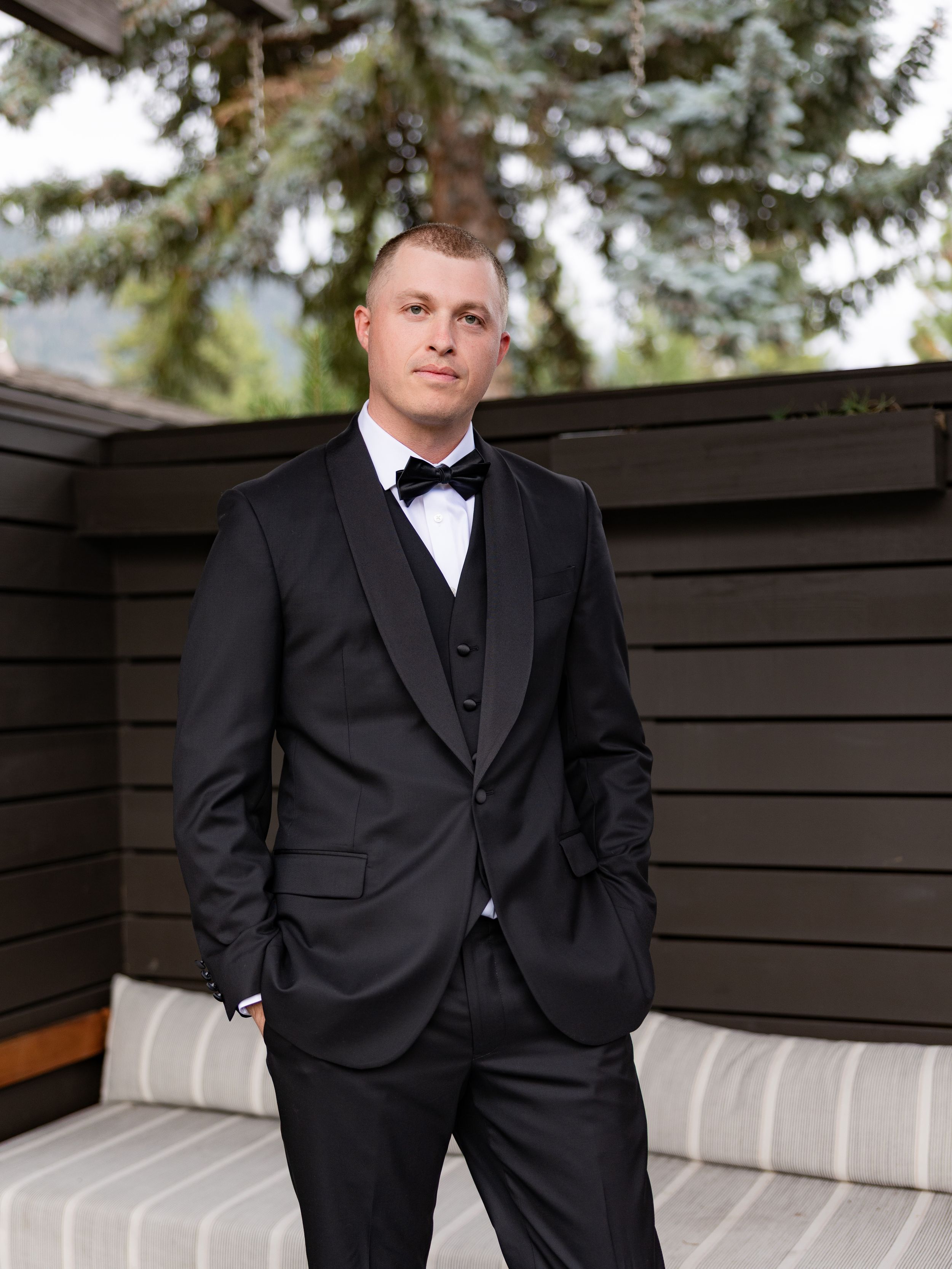 A young man in a black tuxedo with bow tie stands confidently outdoors near wooden fence and evergreen trees.