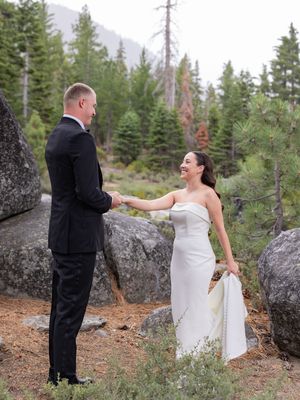 Bride in white strapless gown holds groom's hand outdoors among pine trees and boulders in a mountain wedding setting.