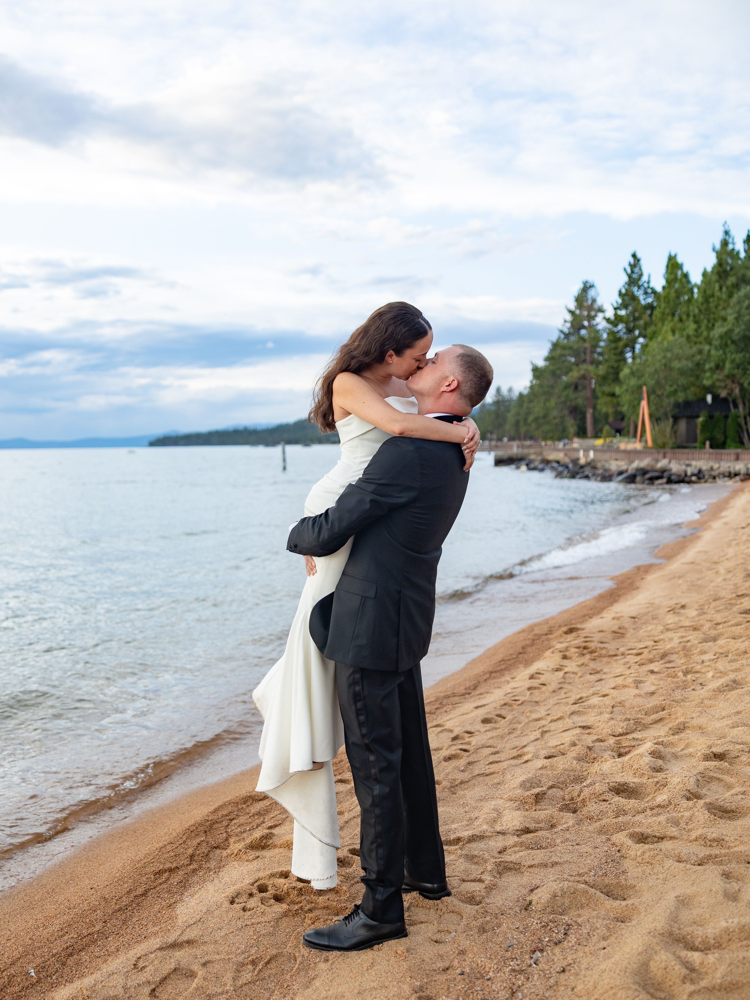 Groom lifts bride in white dress on sandy Lake Tahoe beach shore, sharing a romantic kiss at their wedding.