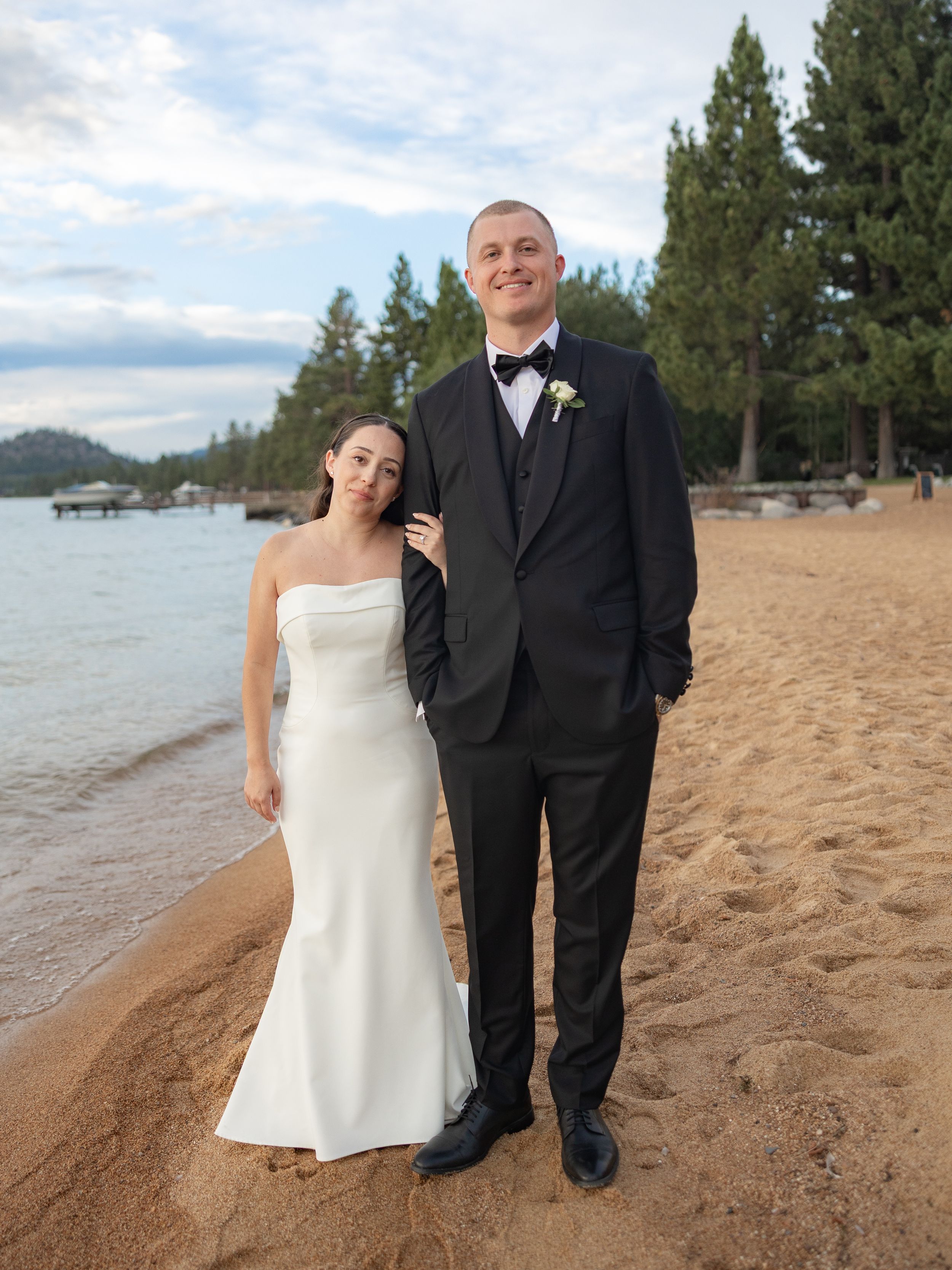 Bride in white strapless gown and groom in black tuxedo posing on sandy Lake Tahoe beach at sunset.