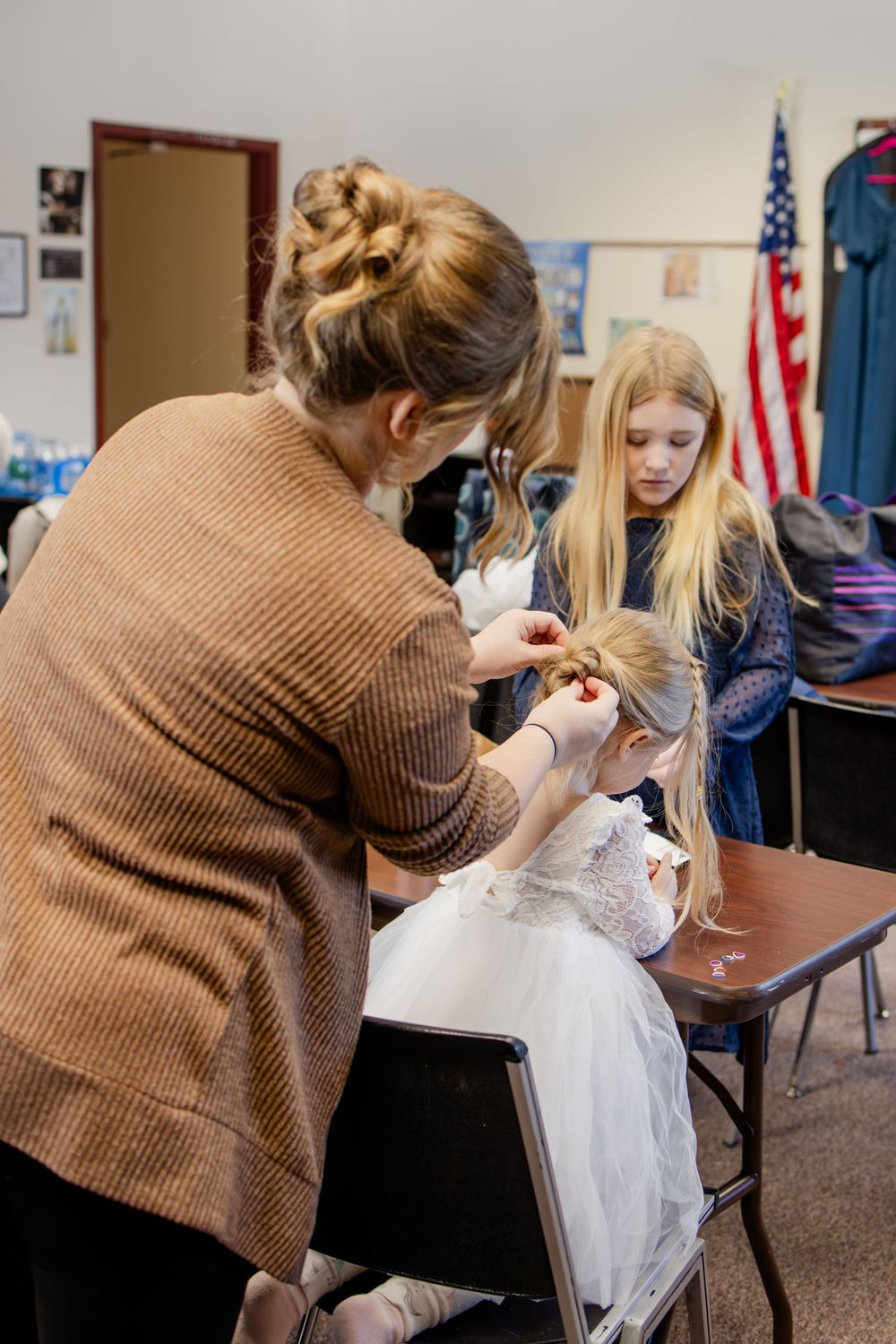 Woman styling young girl's hair in classroom while another blonde girl in blue dress watches near American flag.