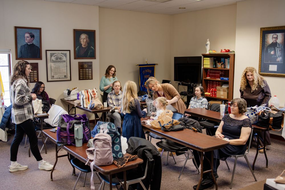 Students gather in a classroom, sitting at desks and socializing, with framed portraits and a bookshelf in the background.