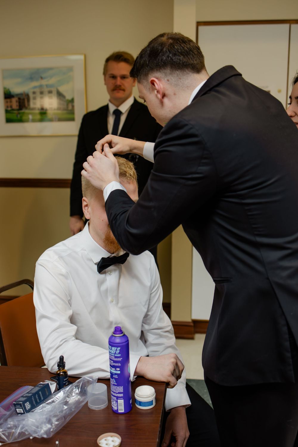 Groomsman styling the groom's hair at a table with hair products before a wedding ceremony.