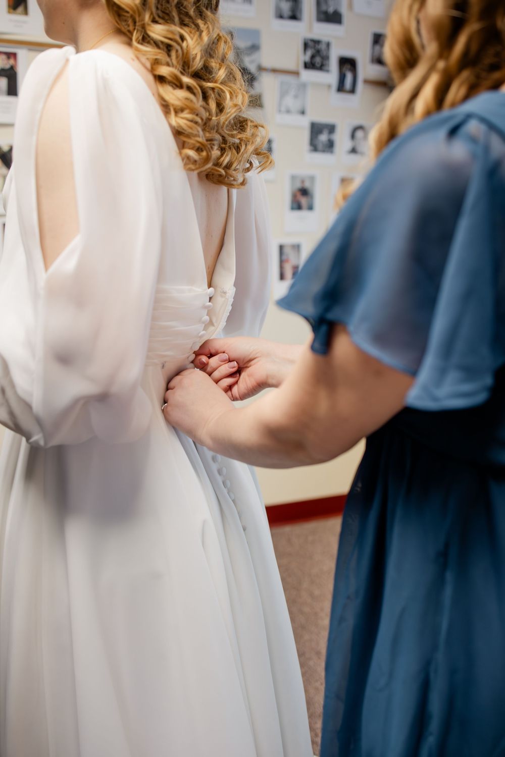 Bridesmaid in blue dress helping bride button up white wedding gown from behind.