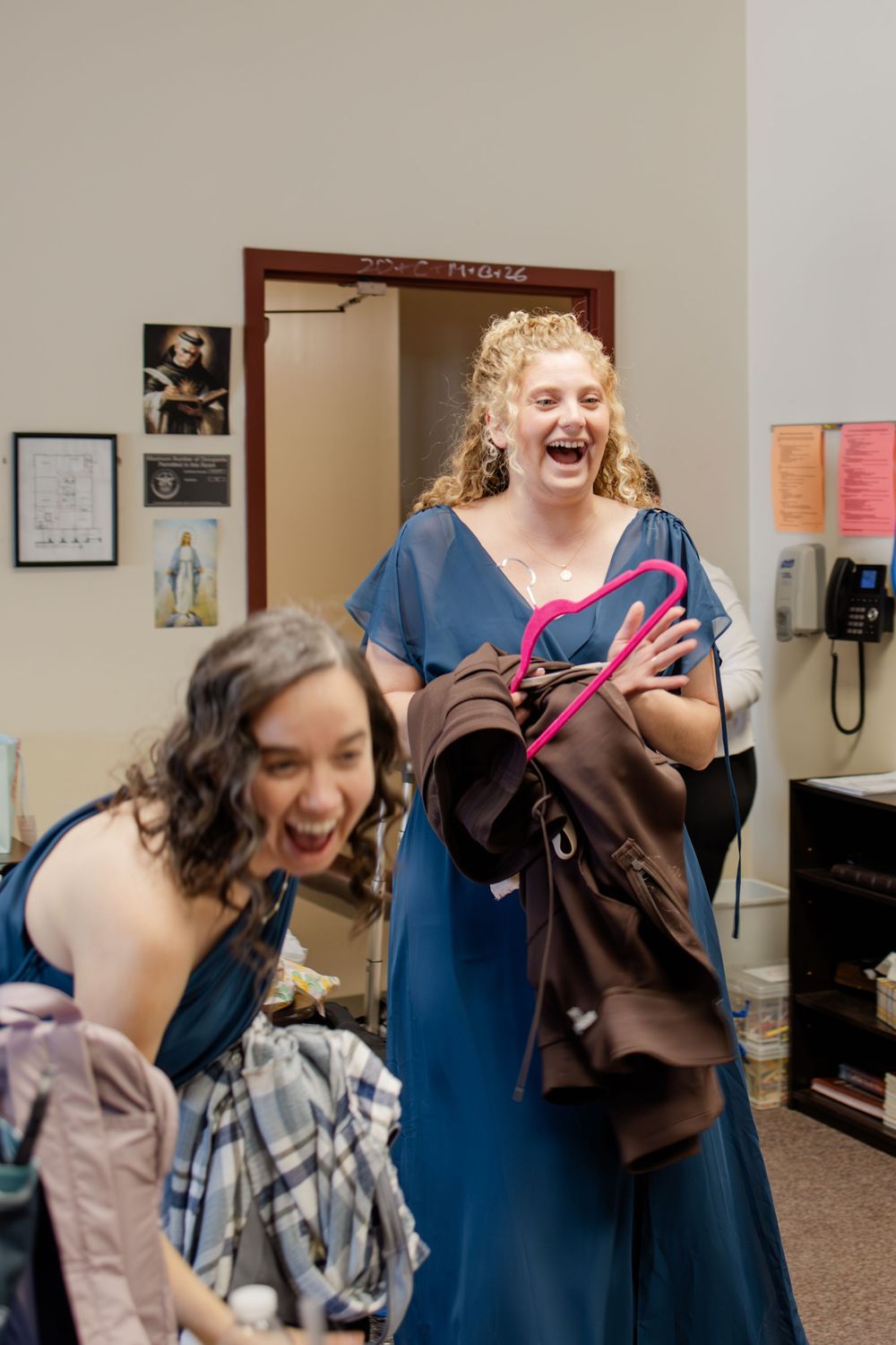 Two women in blue dresses laughing joyfully while holding clothes in a room during wedding preparations.