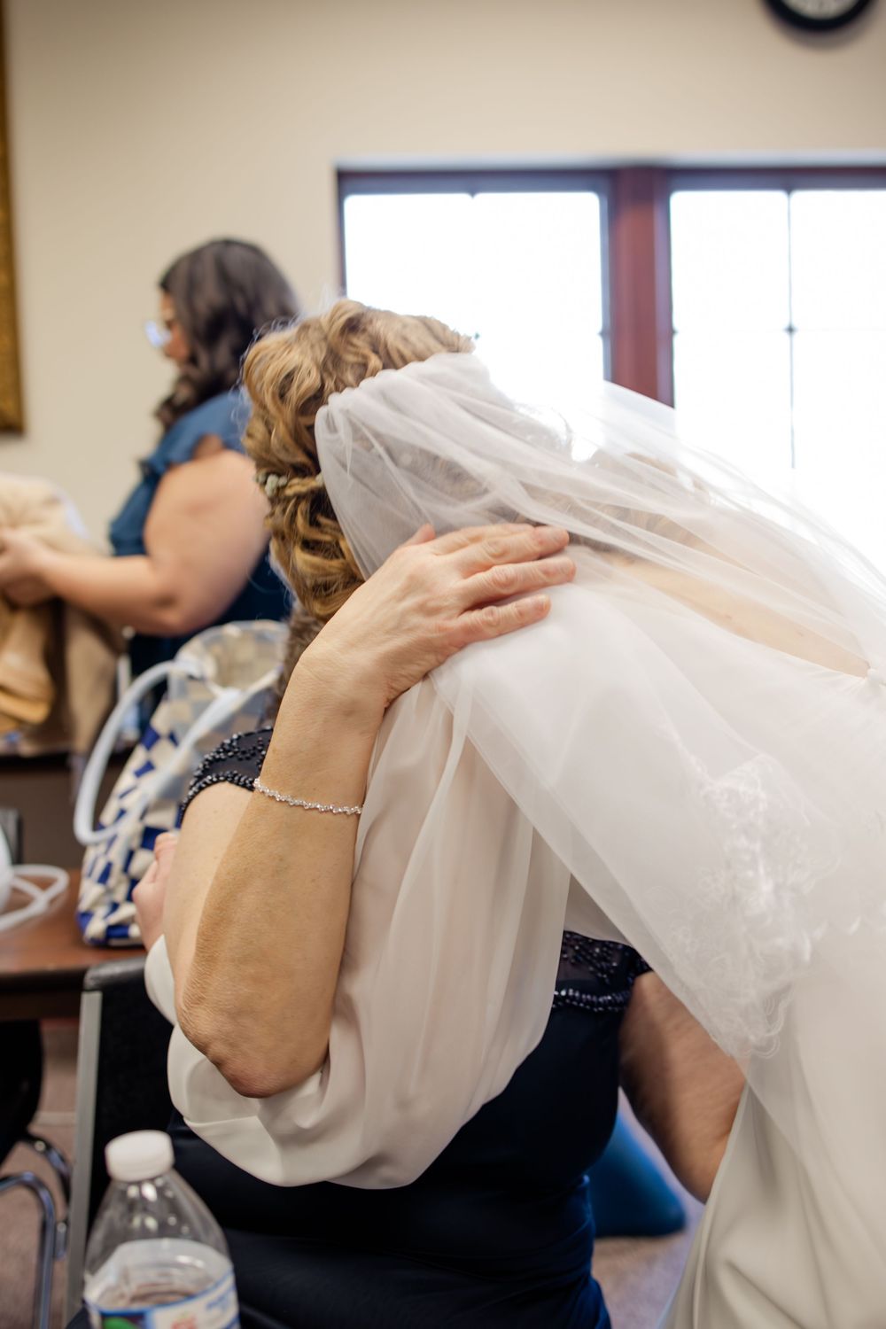 Bride in white veil embracing an older woman in a navy dress during pre-wedding preparations indoors.