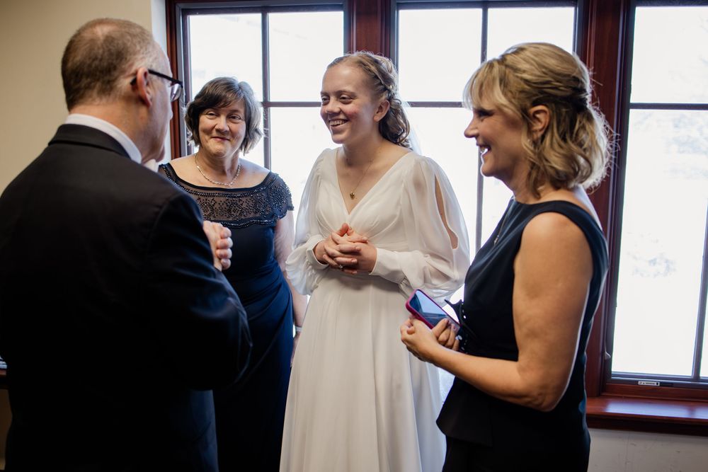 Bride in white gown laughing with wedding guests including a man in a suit and women in navy dresses.