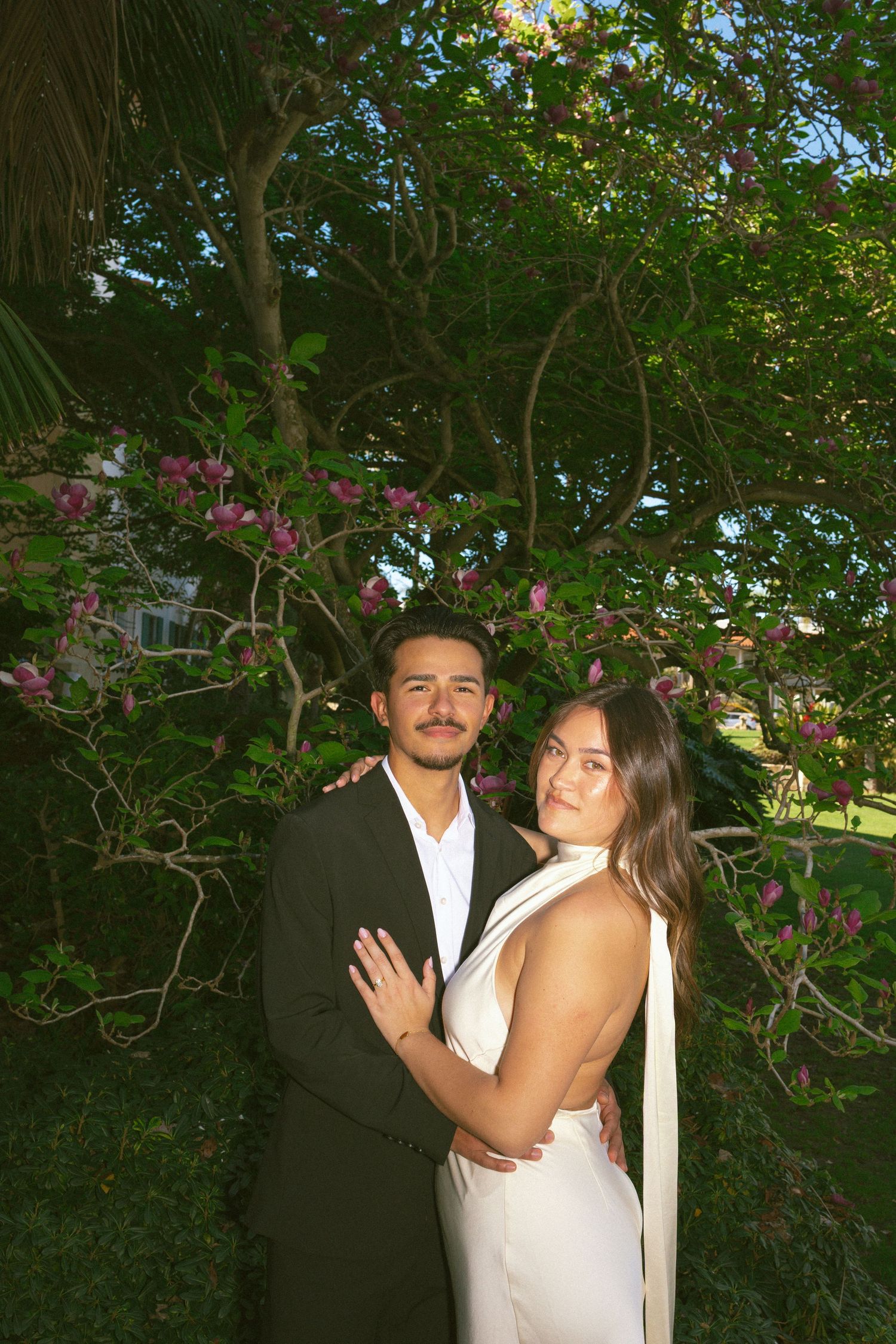 Couple embracing in garden with blooming pink flowers, woman in white dress and man in dark suit.