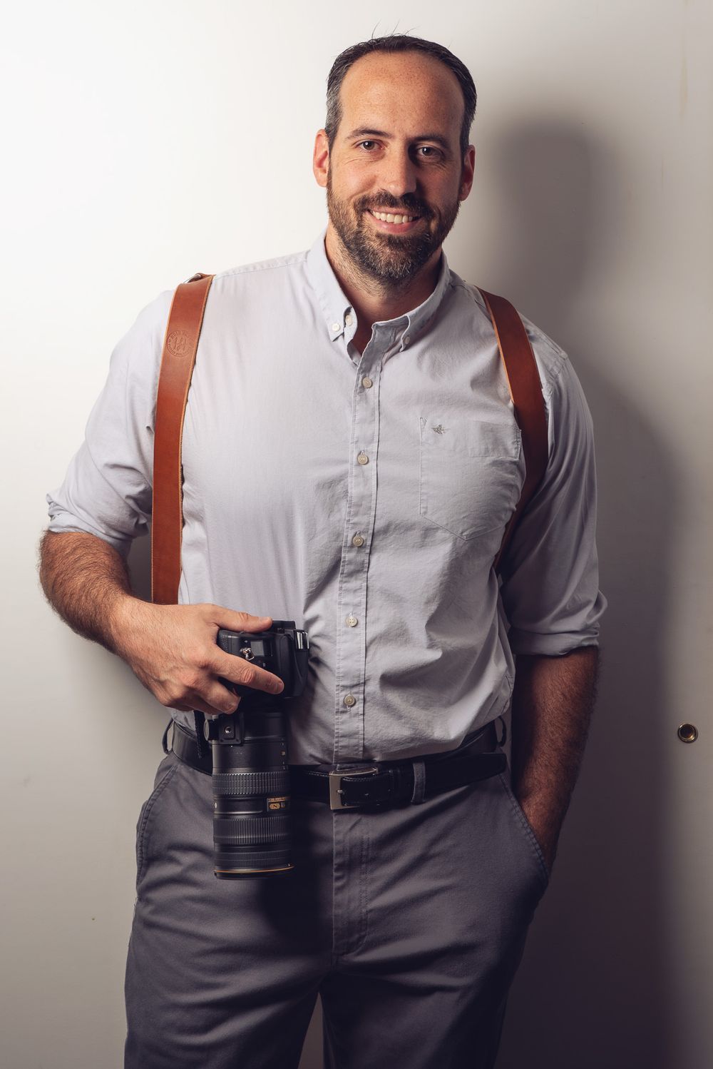Smiling bearded man in gray shirt holding a DSLR camera with brown leather straps, posing against a white wall.