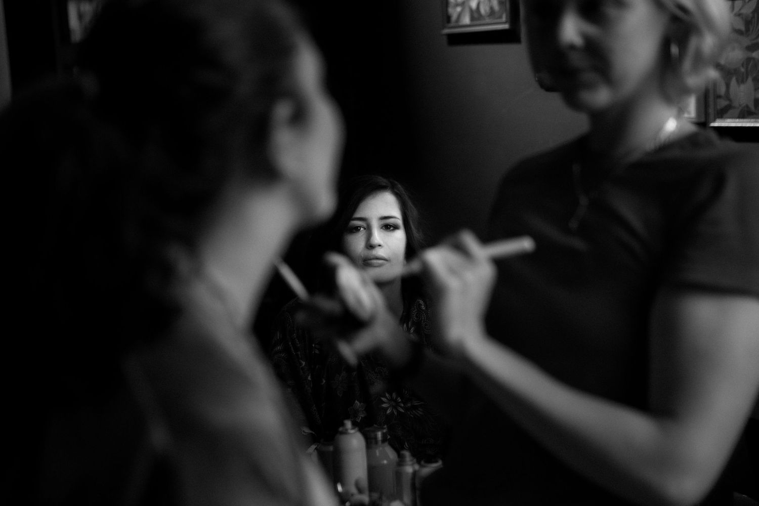 Black and white photo of a woman getting makeup applied by a makeup artist, with her reflection visible in a mirror.