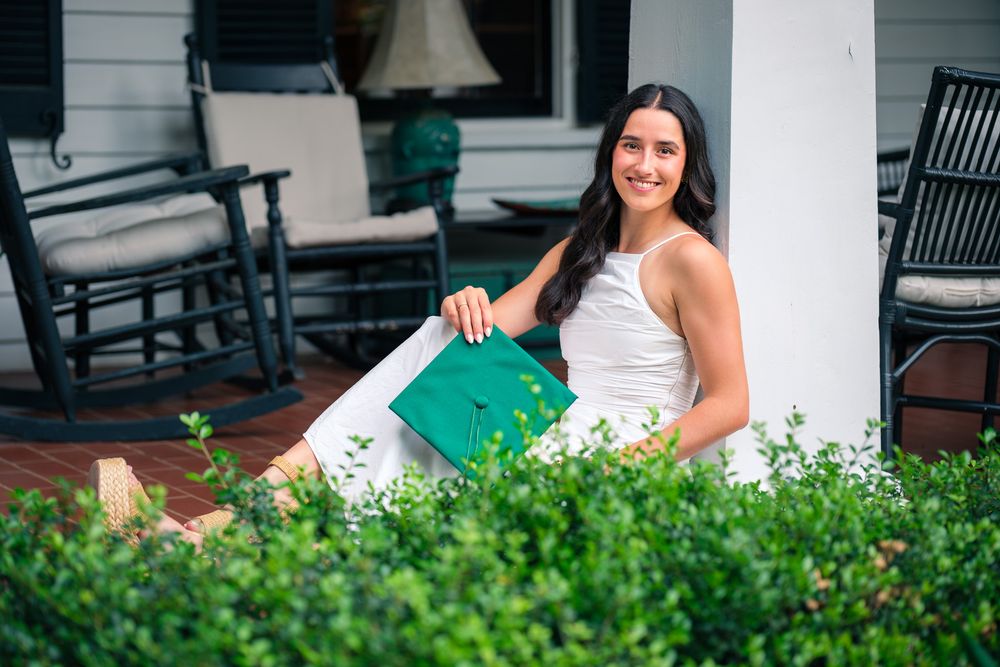 Senior cap and gown session photo session with smiling female graduate sitting on a porch holding a green graduation cap outdoors.