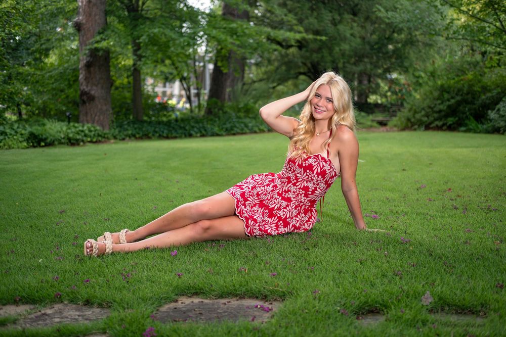 Blonde woman in red floral sundress sitting on green grass in a park, posing with hand in hair.