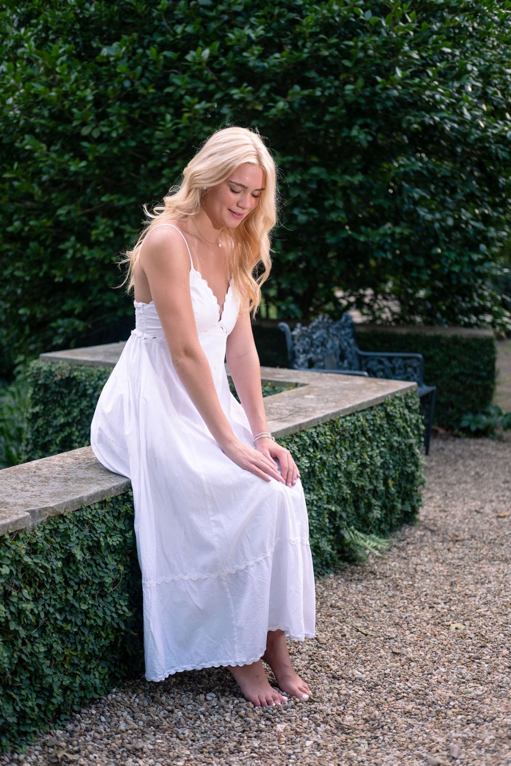 Blonde woman in white sundress sitting barefoot on stone garden wall surrounded by lush green hedges.