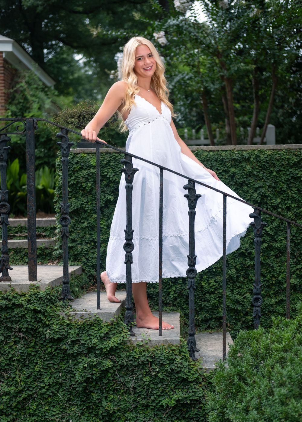 Blonde woman in flowing white midi dress posing on stone steps with black iron railing surrounded by lush green hedges.