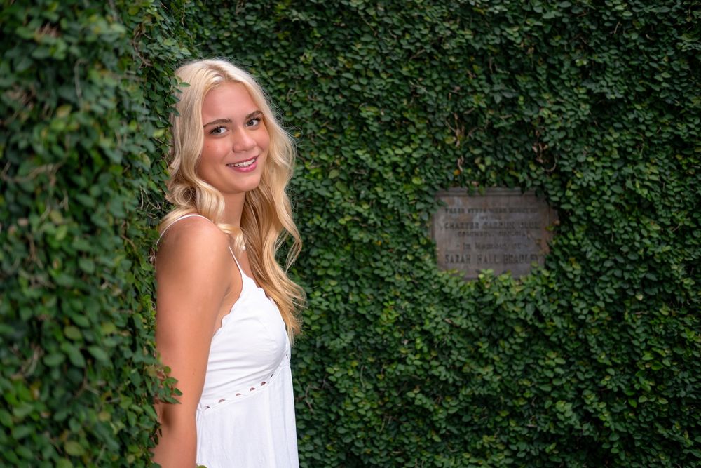 Young blonde woman in white dress smiling in front of lush green ivy wall with decorative plaque.