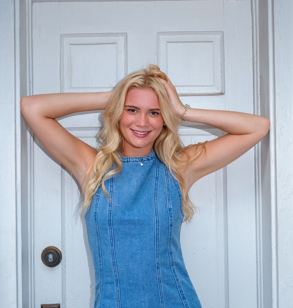 Smiling blonde woman in a denim dress posing with hands behind her head in front of a white door.