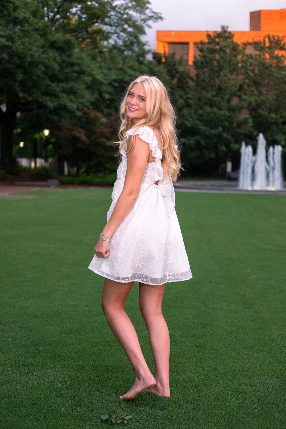 Young blonde woman in white lace dress standing barefoot on green grass near fountain.