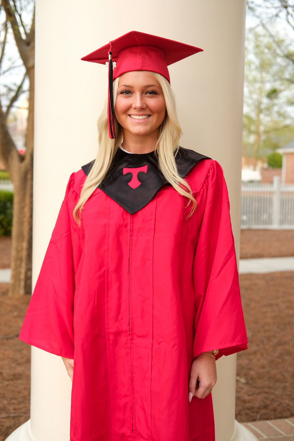 Smiling blonde woman in red graduation cap and gown with black collar standing by a white column outdoors.