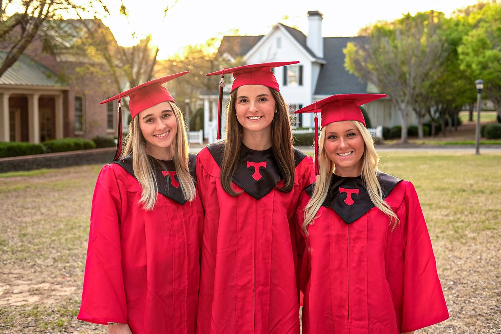 Three smiling women in red graduation caps and gowns pose outdoors near a house on a sunny day.