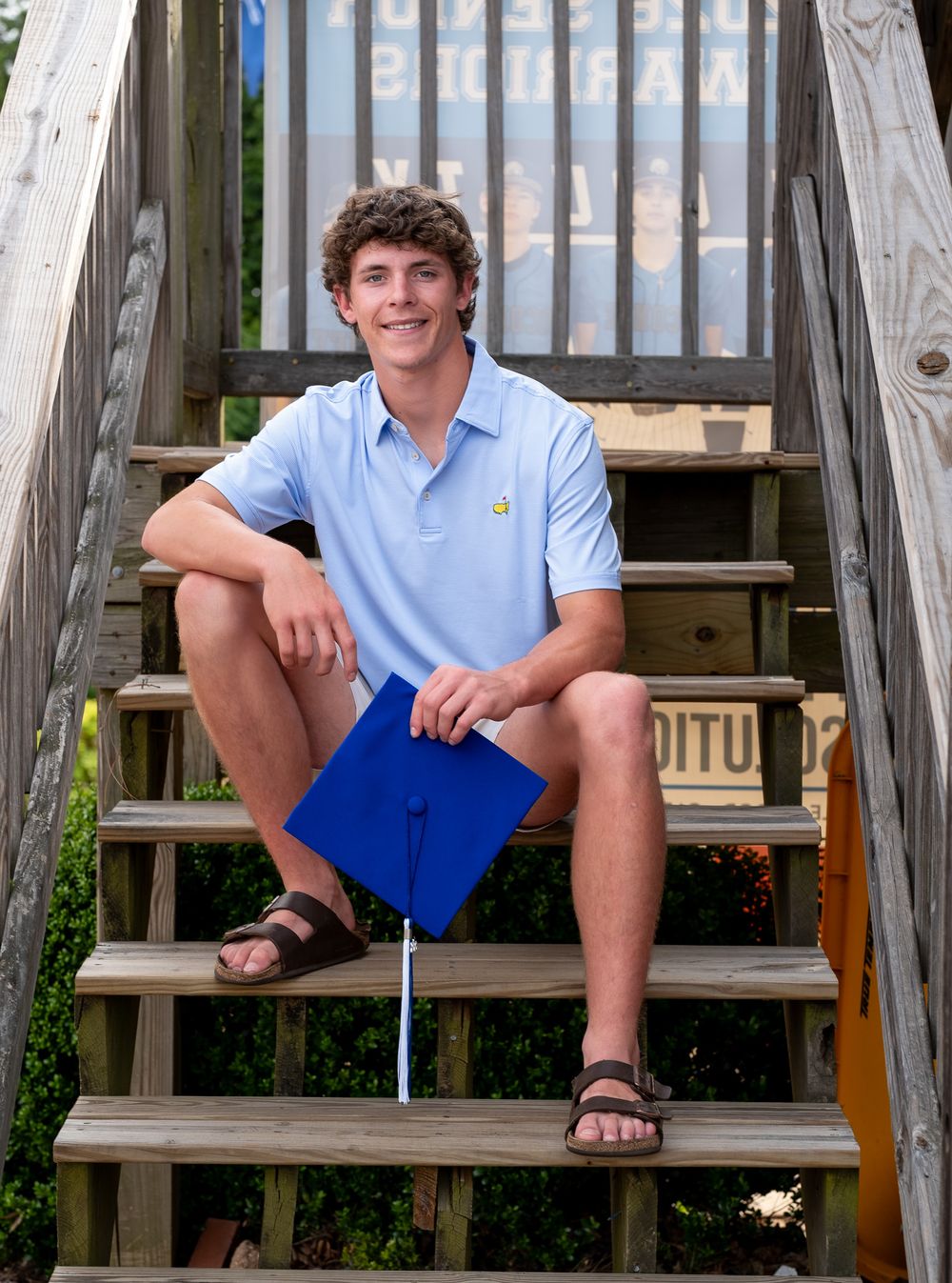 Smiling senior graduate sitting on wooden steps holding a blue graduation cap outdoors for his senior cap and gown photoshoot.
