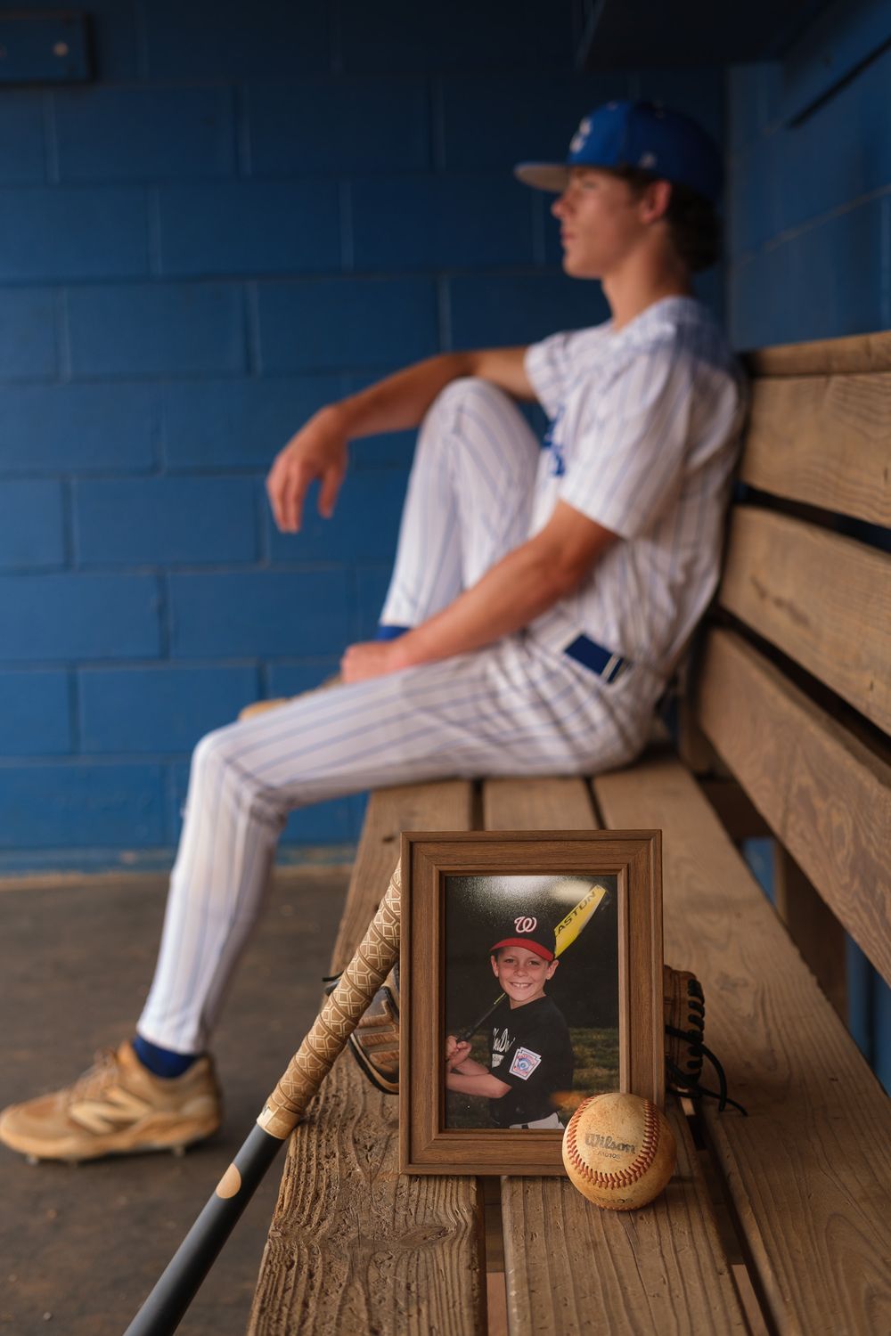 Baseball player sitting in dugout beside a framed childhood photo, bat, and ball for his senior cap and gown photoshoot.