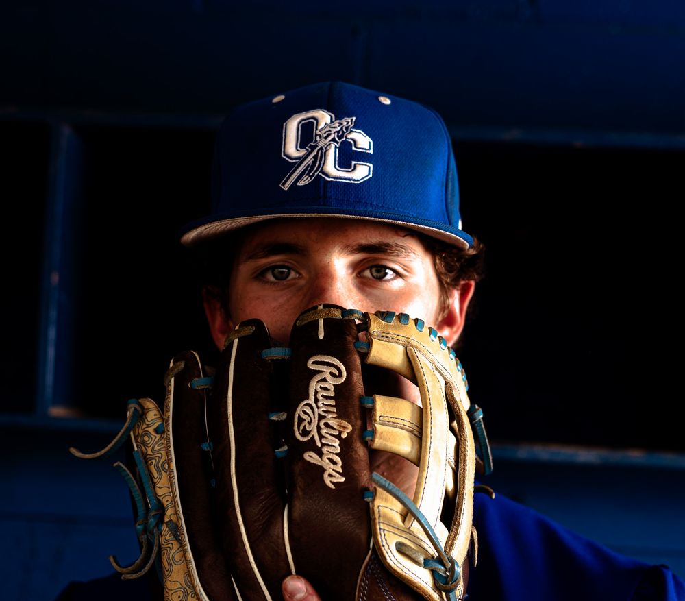 Senior photoshoot with a Baseball player covering face with a Rawlings glove wearing a blue OC team cap.