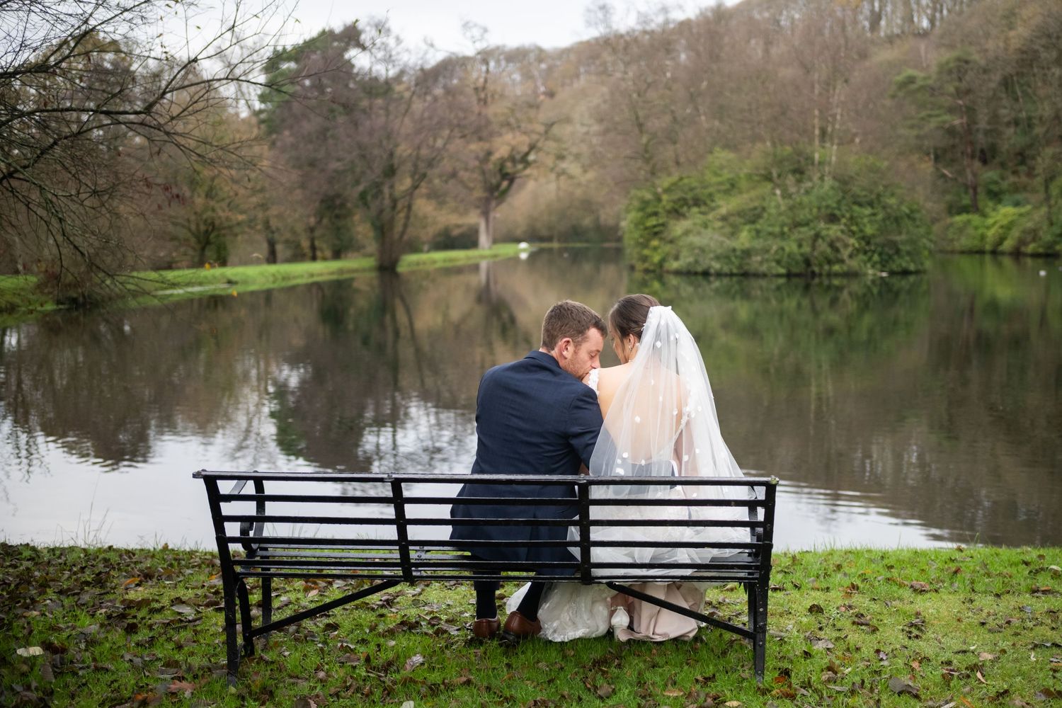 Newlywed couple sitting on a bench by a serene lake, bride in white gown and veil, sharing a romantic moment outdoors.