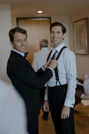 Two people in formal attire help each other get dressed before a special event in a warmly lit room.